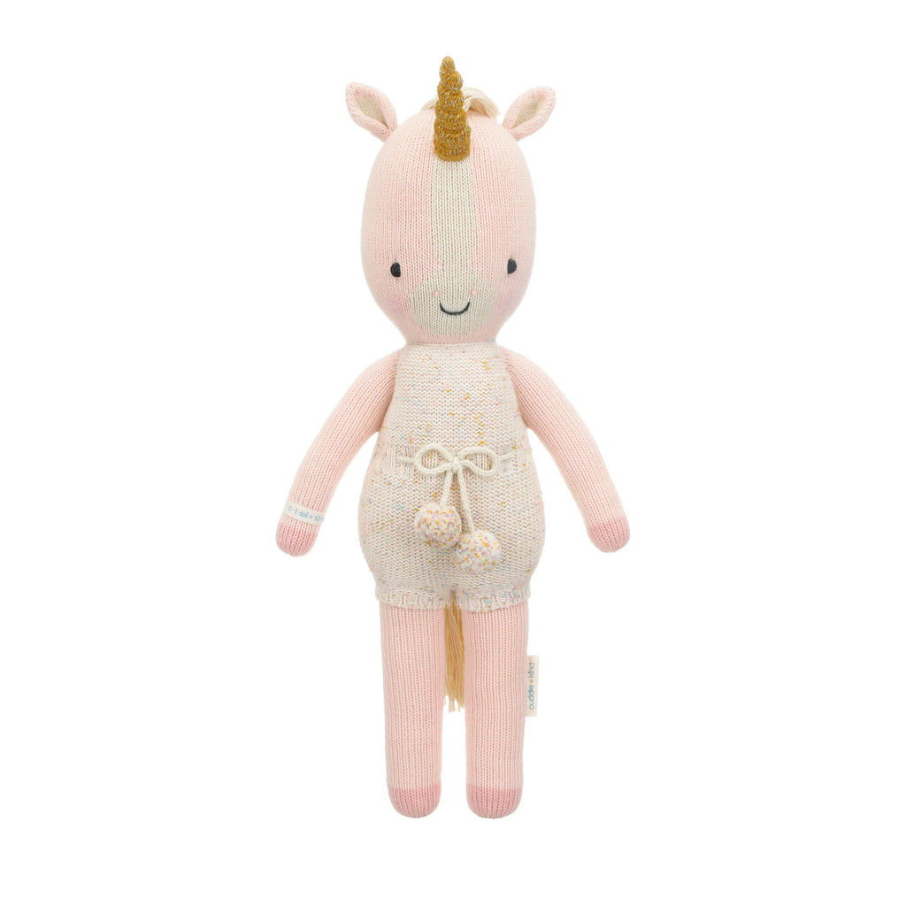 Cuddle + Kind Ella the Unicorn is a plush best friend doll with a pink body, cream horn, cream overalls with pompoms, and embroidered eyes. Each hand-knit doll helps provide meals to children in need.