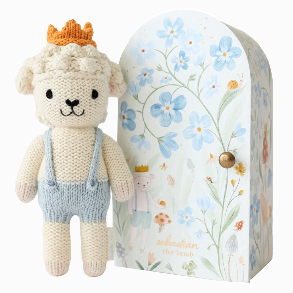 A handmade Cuddle + Kind Tiny Sebastian the Lamb, wearing a crown and blue overalls, stands by a decorative box with flower illustrations and another crowned lamb. The box reads "Sebastian the Lamb," and this charming piece is crafted with fair trade principles.