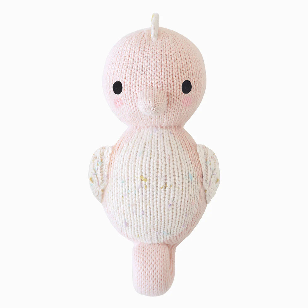 The Cuddle + Kind Baby Seahorse in Powder Pink features a charming round body, tiny beak, and small wings. Hand-knit from natural Peruvian cotton yarn with black eyes on a white backdrop, this delightful collectible promises to enchant.