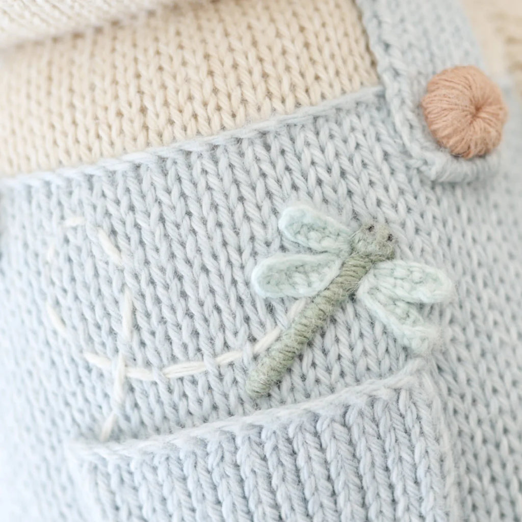Close-up of light blue knit fabric with a small embroidered dragonfly and white stitched line; a beige button and cream knitting from the fair trade, hand-knit Cuddle + Kind Bailey The Mouse doll appear in the background.