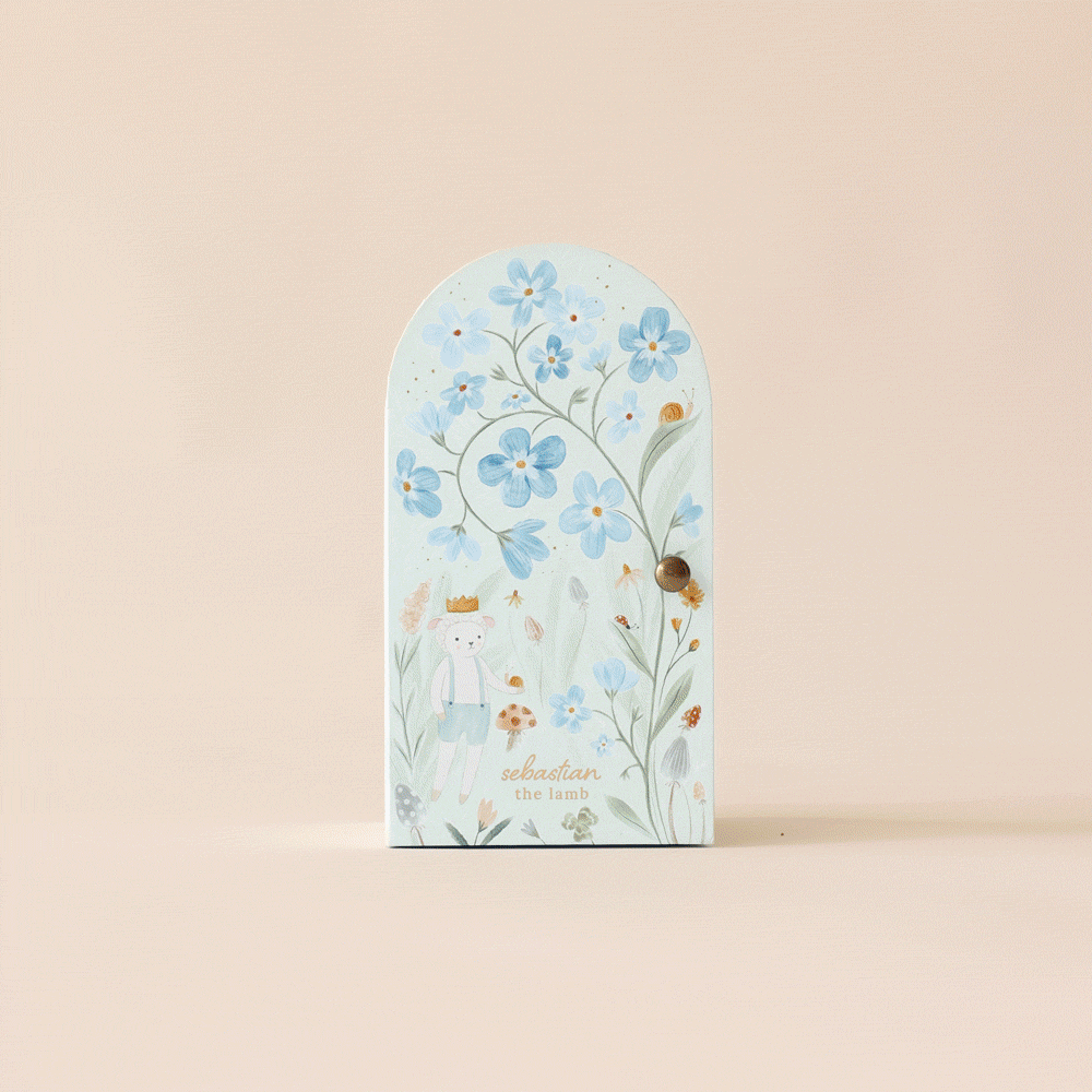 A sustainably handmade, arch-shaped card features blue flowers, a crowned illustrated lamb named Sebastian, and various insects on a pastel pink background. The text "Sebastian the lamb" appears in light green. Product: Cuddle + Kind Tiny Sebastian the Lamb.