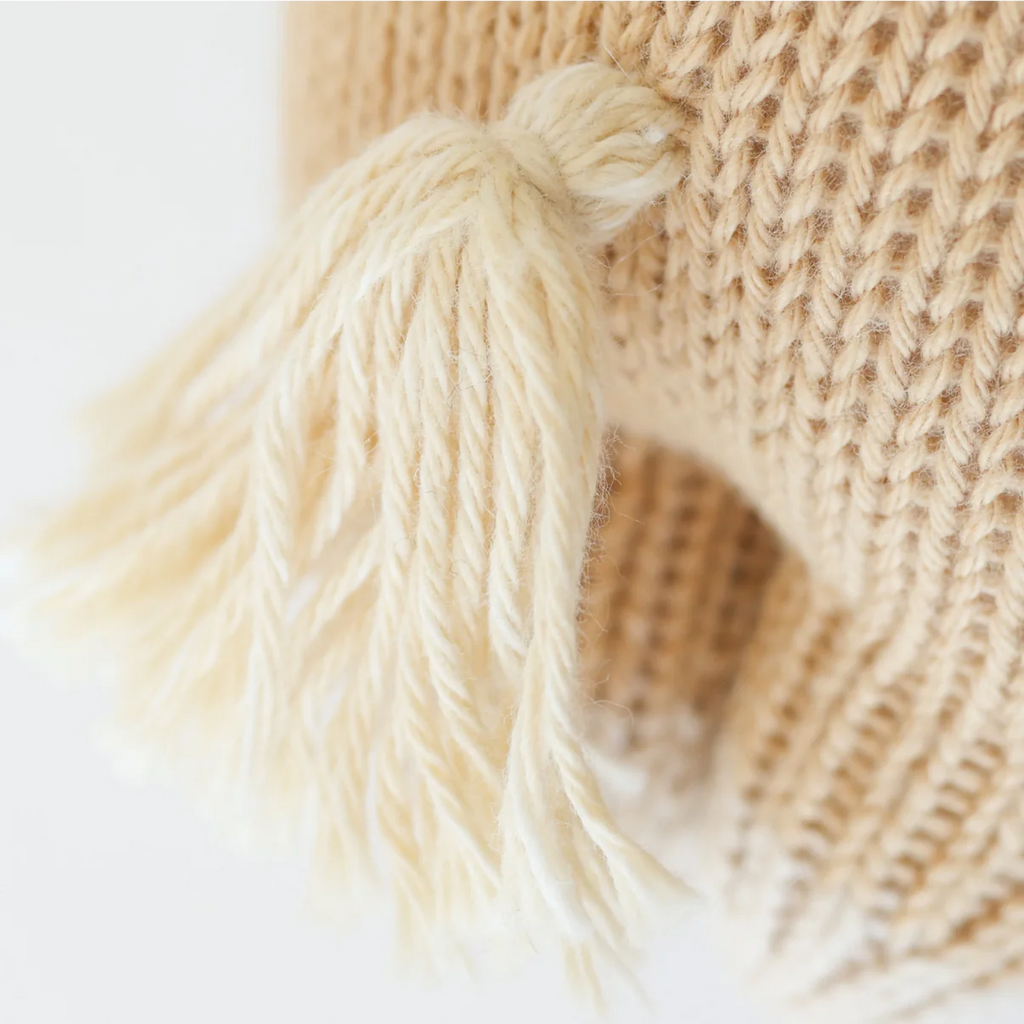 Close-up of the Cuddle + Kind Baby Pony’s beige knitted Peruvian cotton fabric with a cream tassel, highlighting detailed stitches and fibers, all crafted using sustainable fair trade practices.