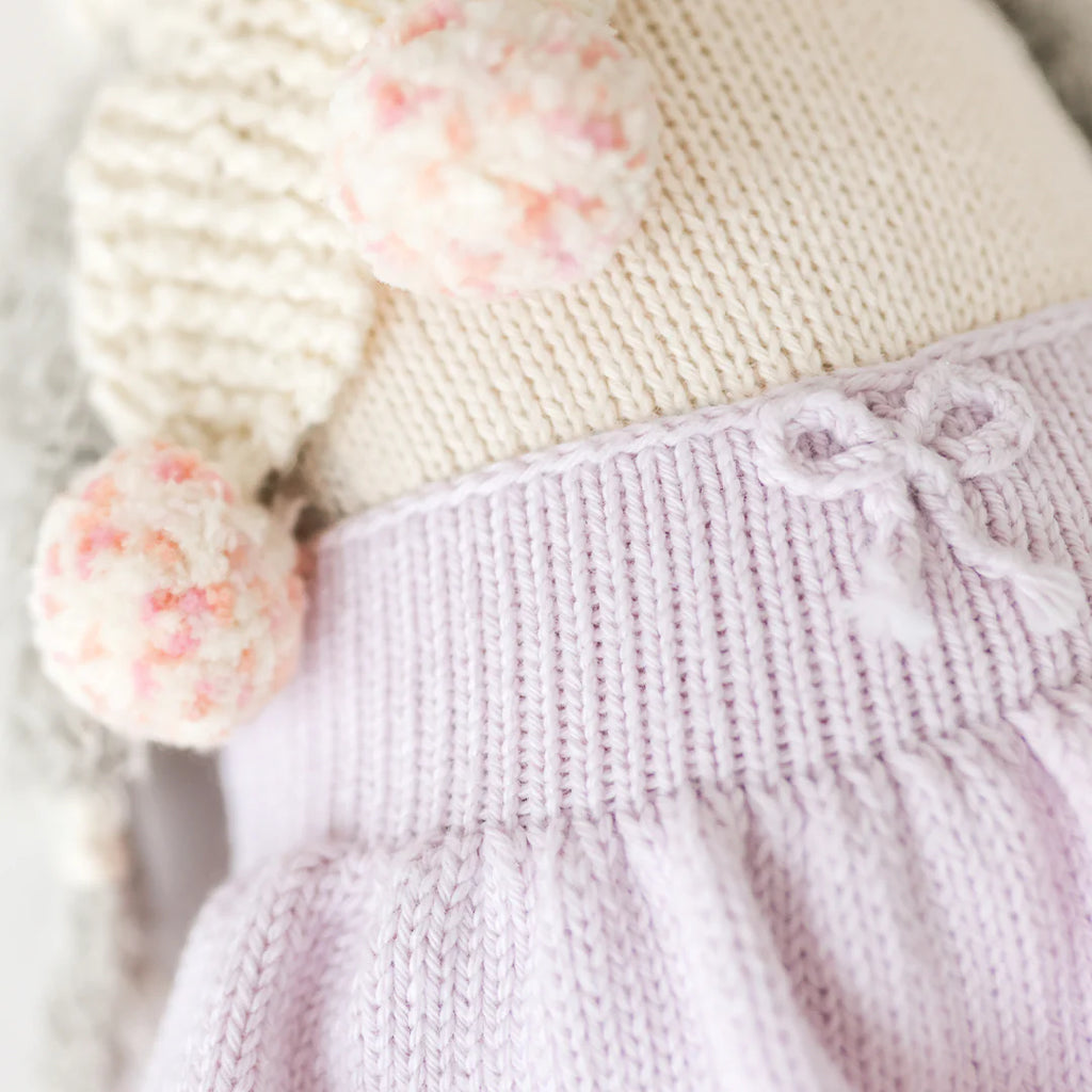 Close-up of Cuddle + Kind Skylar The Penguin, a fair trade knit cotton doll dressed in a pastel lilac skirt with a bow at the waistband, cream top, and soft pink and white pom-pom decorations.