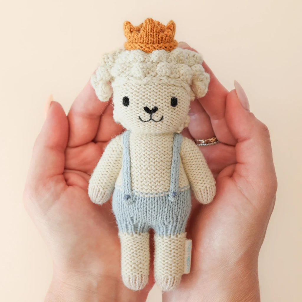 Held gently by two hands, Cuddle + Kind's tenderly knitted Tiny Sebastian the Lamb sports a charming smile with cream-colored fur and wears light blue overalls and a small brown crown, all set against a plain, light beige background.