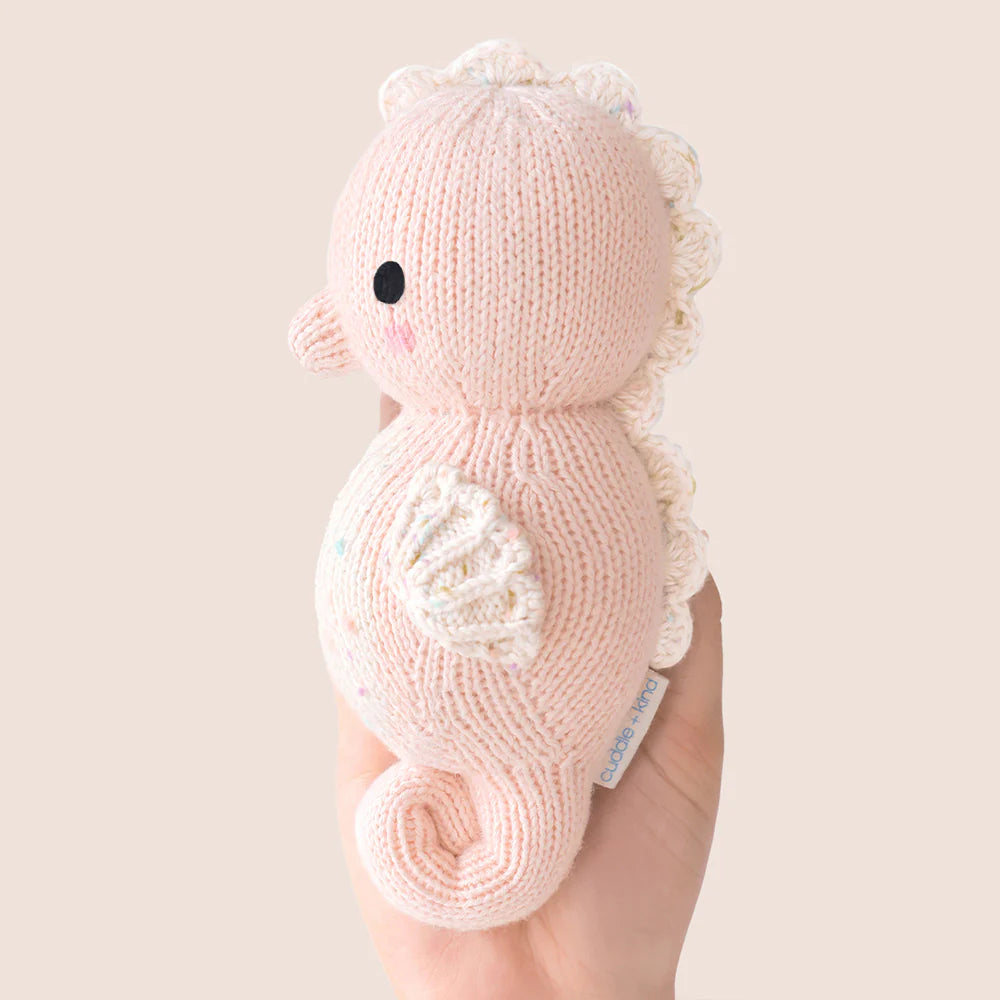 Someone holds the Cuddle + Kind Baby Seahorse (Powder Pink) plush toy, hand-knit from natural Peruvian cotton. It features a scalloped white mane, small wings, and colorful spots against a plain beige background—a delightful collectible for baby animal fans.
