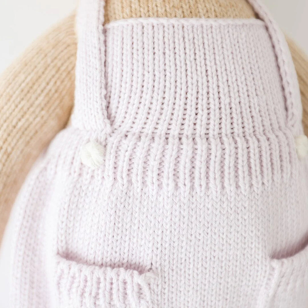 A close-up of a beige knitted Cuddle + Kind the Honey Bear wearing light pink, hand-knitted overalls. The overalls feature small pockets and are detailed with ribbed edges and simple knitted straps. The soft texture and hypoallergenic polyfill give a warm, cozy, and handmade feel.