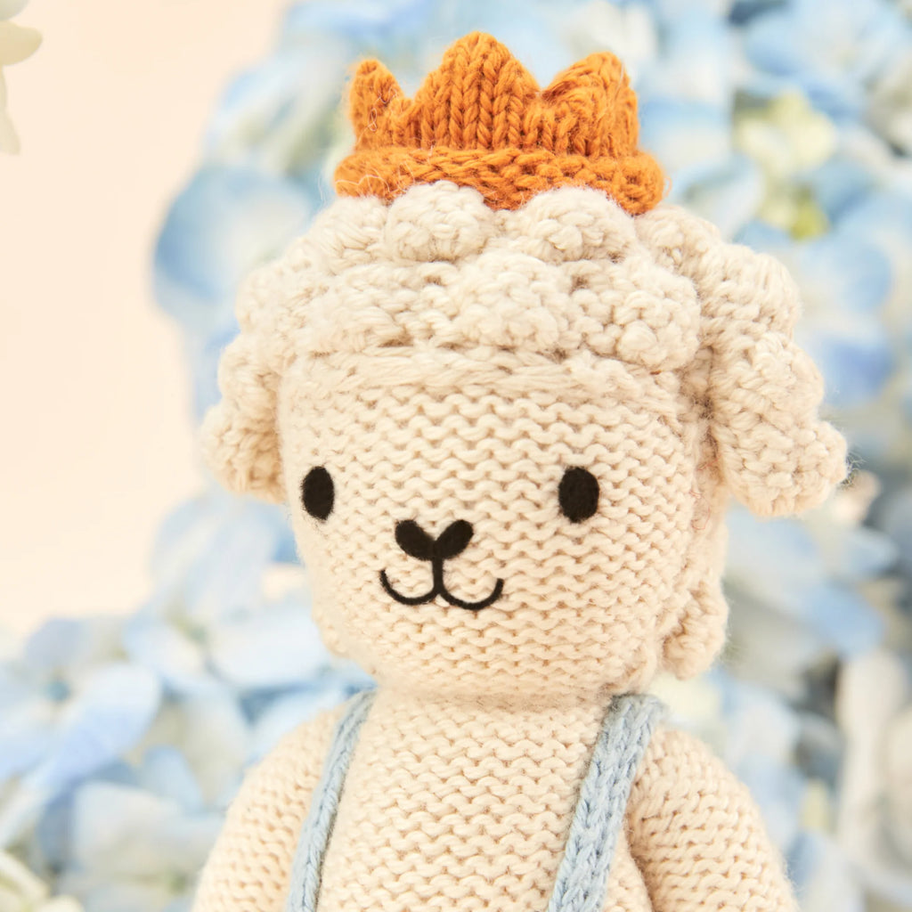 Cuddle + Kind's Tiny Sebastian the Lamb is a crocheted lamb toy with an orange crown and light blue overalls, made from sustainable materials, pictured against a blurred backdrop of blue flowers.