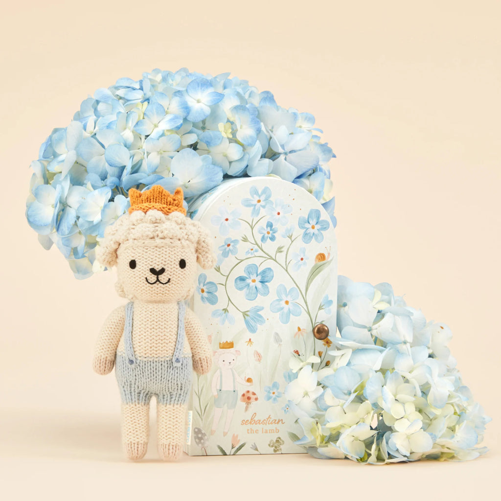 The Cuddle + Kind Tiny Sebastian the Lamb, dressed in blue overalls and a crown, stands next to a decorative box with blue flowers and cascading hydrangeas, creating a charming pastel scene that celebrates fair trade artistry.