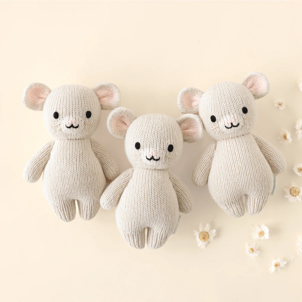 Three cream-colored Cuddle + Kind Baby Mouse plush toys with round ears, black eyes, and small smiles are placed side by side on a beige background with white flowers. Handmade in Peru and Nepal from Peruvian cotton yarn.
