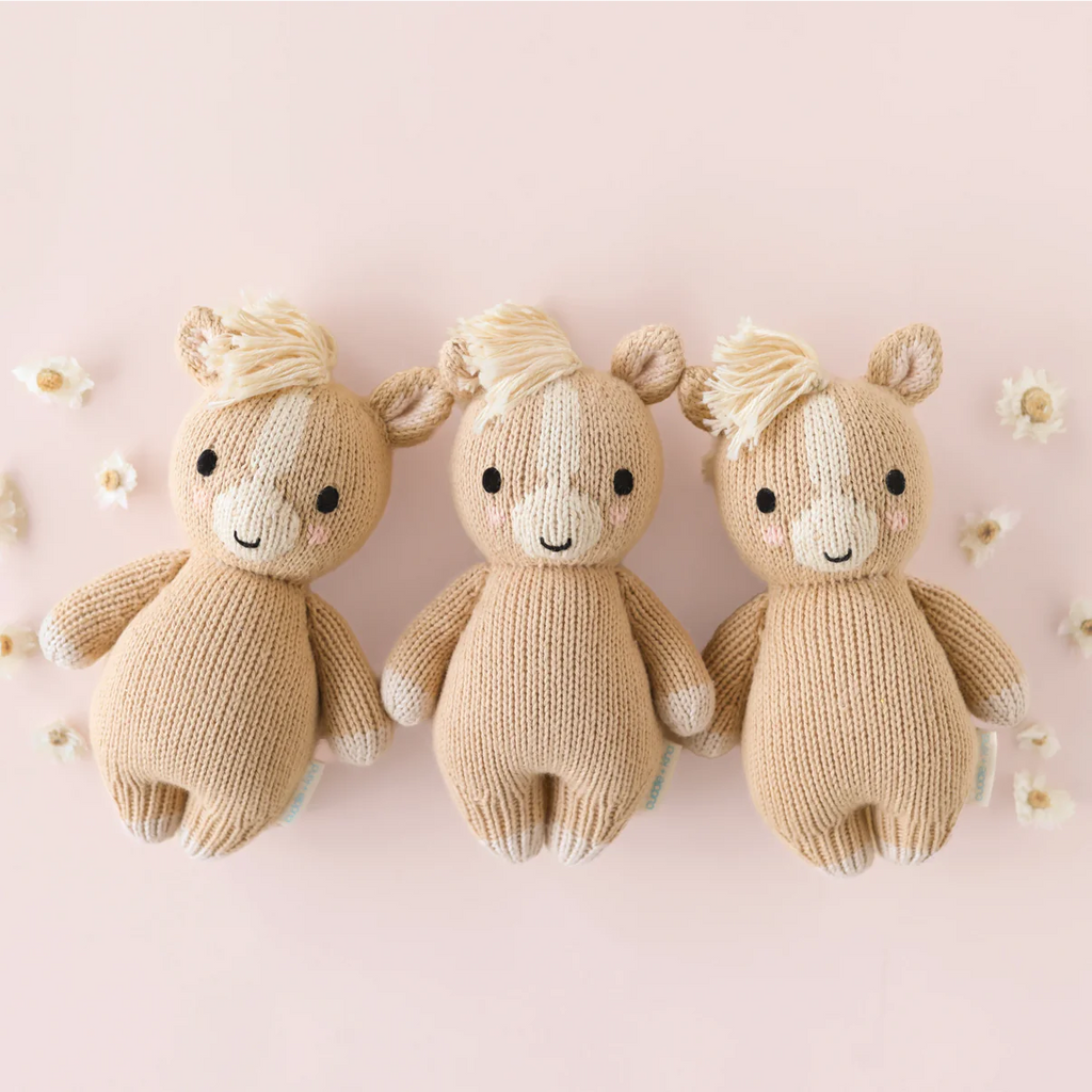 Three Cuddle + Kind Baby Pony plush toys—beige with white manes and tails—are hand-knit from soft Peruvian cotton yarn, arranged side by side on a pale pink background surrounded by small white flowers.