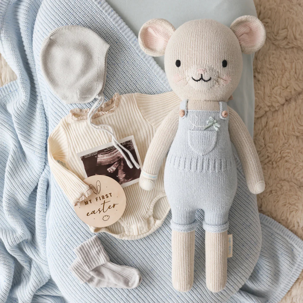 A soft blue blanket, cream-colored baby onesie with lace collar, gray bonnet and socks, a "My first Easter" tag, and a large Cuddle + Kind Bailey The Mouse doll in blue overalls are arranged on a beige textured surface.