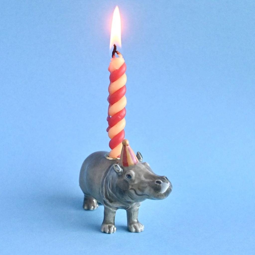 A whimsical image of a small, gray, hand-painted Hippo Cake Topper with a twisted, lit orange candle on its back against a plain light blue background.