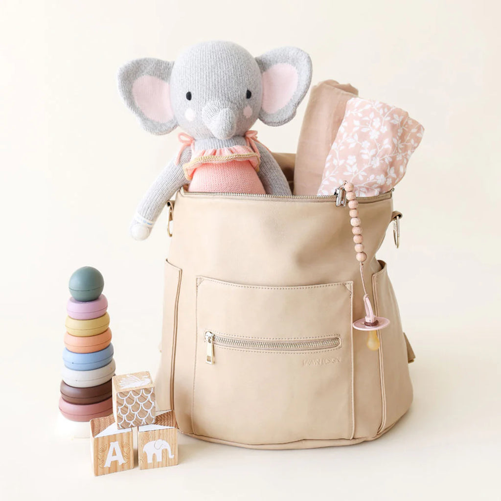A beige diaper bag with a Cuddle + Kind Eloise the Elephant, a pink floral blanket, and a pacifier hangs on the side. In front are colorful stacking rings and wooden blocks with animal and letter designs.