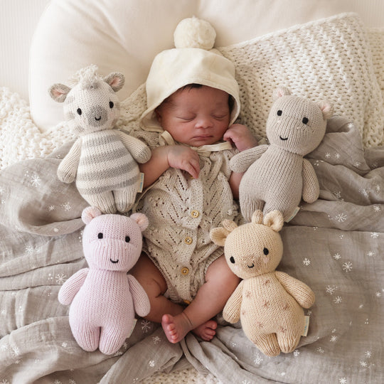 Newborn baby sleeping peacefully surrounded by four Cuddle + Kind Baby Giraffe toys, wearing a knitted hat and wrapped in a cozy blanket.