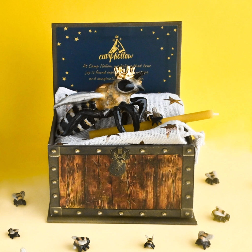 A Queen Bee Cake Topper styled like a treasure chest with a spider and web motif, accompanied by a book titled "Camp Hollow." The background is yellow with scattered small spider figures, and the cake topper is hand painted.