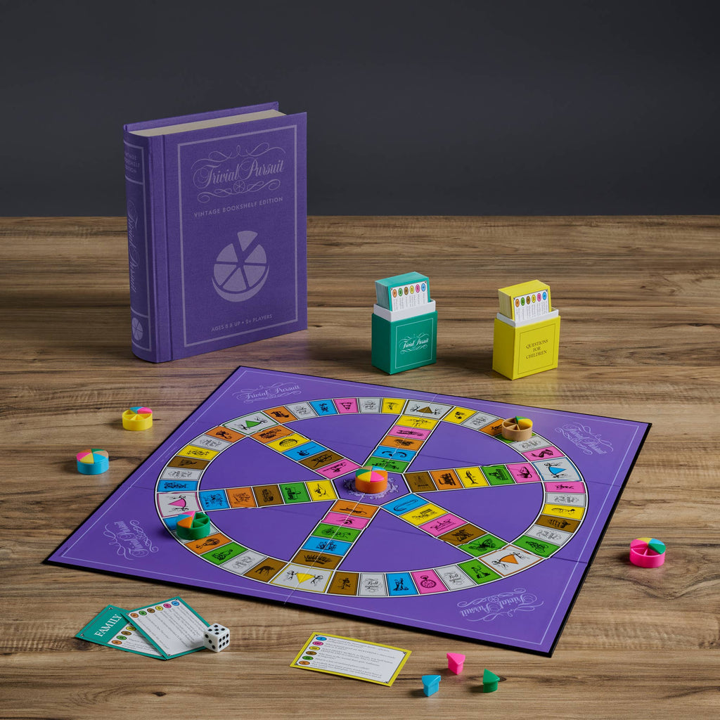The Board Game: Trivial Pursuit Vintage Bookshelf Edition is displayed on a wooden table with colorful pieces, two card boxes, a die, question cards, and its large purple book-style box in the background.