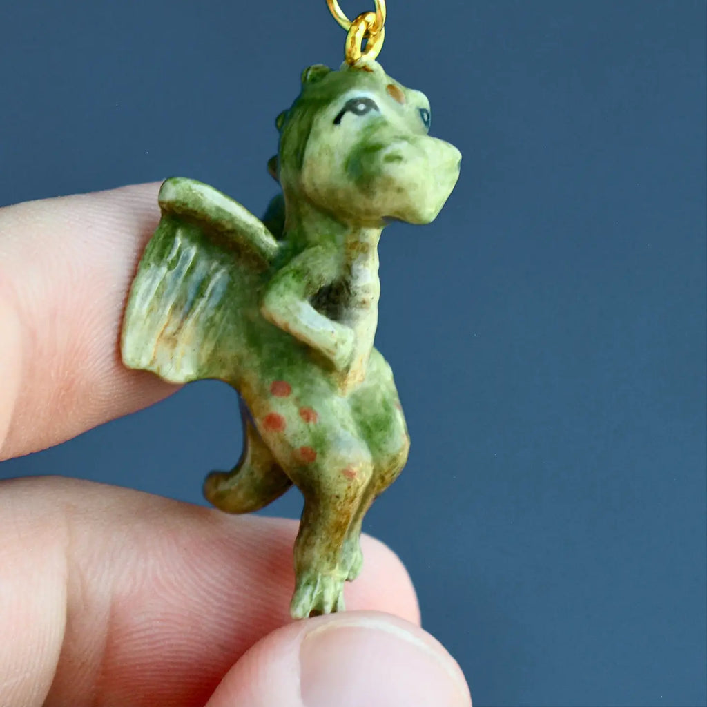 A person holds a small, whimsical handcrafted Baby Dragon Necklace against a grey background. The pendant has a loop on top for hanging.