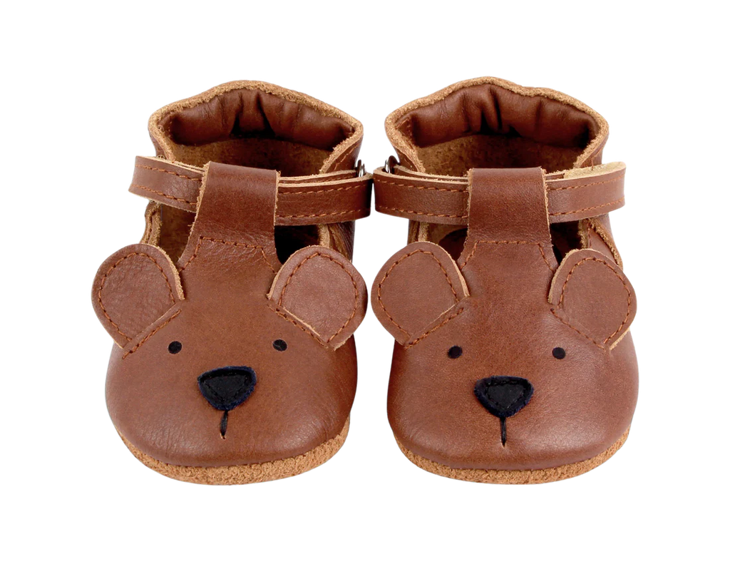 A pair of Donsje Baby Bear Shoes made from premium leather, designed to look like bears, featuring rounded ears, stitched eyes, and black noses with a Velcro fastening strap, isolated on a black background.