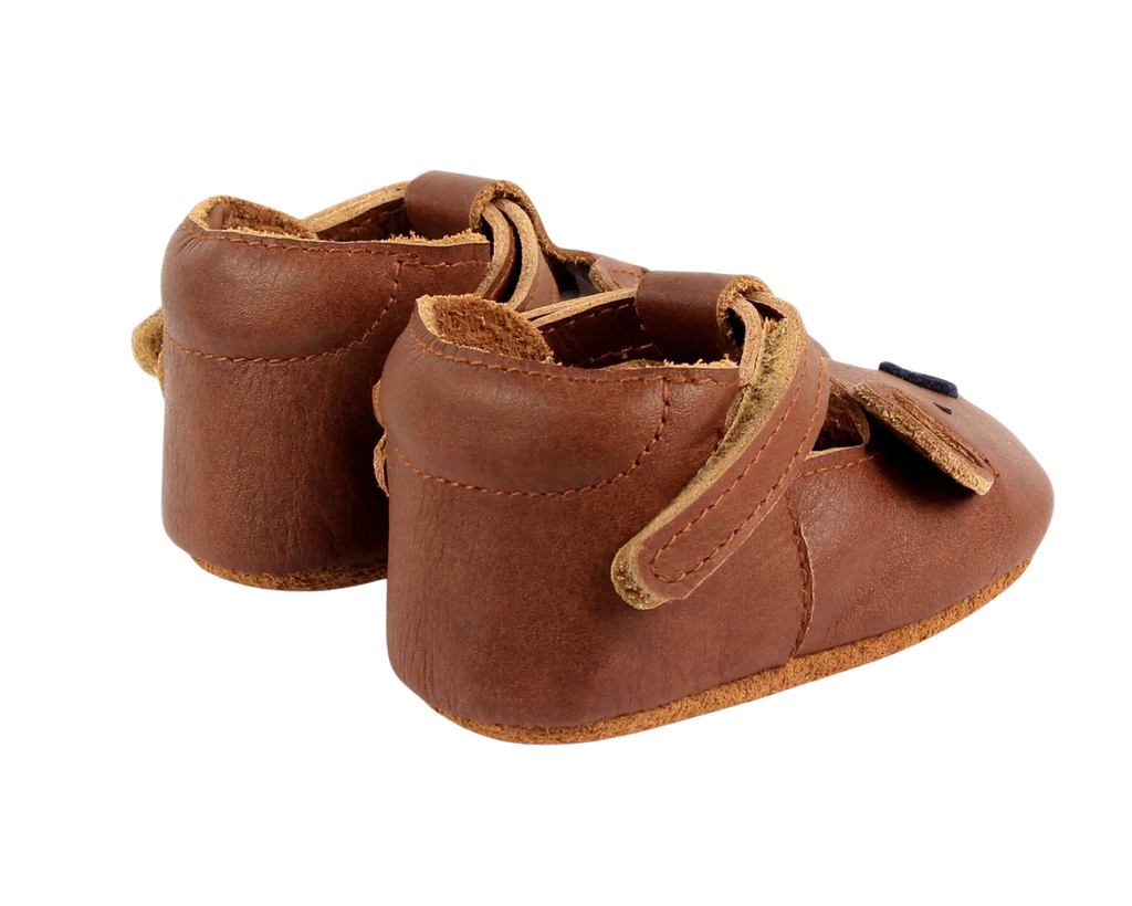 A pair of small, brown Donsje Baby Bear Shoes with Velcro fastening straps, displayed against a black background.