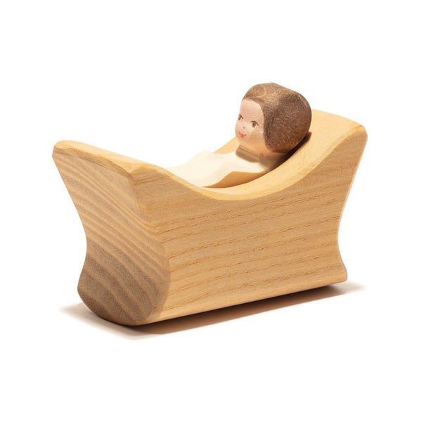Ostheimer Child In Cradle. The baby has a painted face, brown hair, and is wrapped in a light-colored swaddle. The cradle, reminiscent of Ostheimer toys, features a minimalist, curved design with visible wood grain patterns that invite imaginative play.