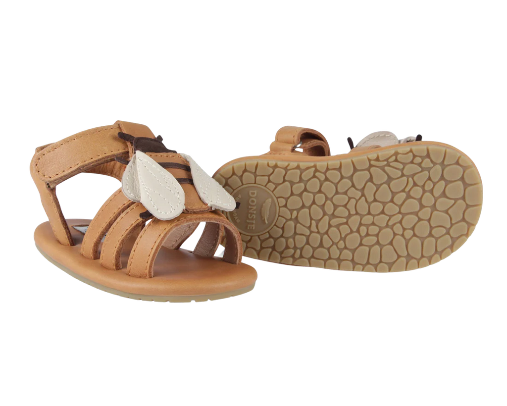 A pair of Donsje Baby Bee Shoes with tan straps, white petal-shaped embellishments, and a non-slip tan sole featuring a Velcro closure. The shoes are displayed against a white background.