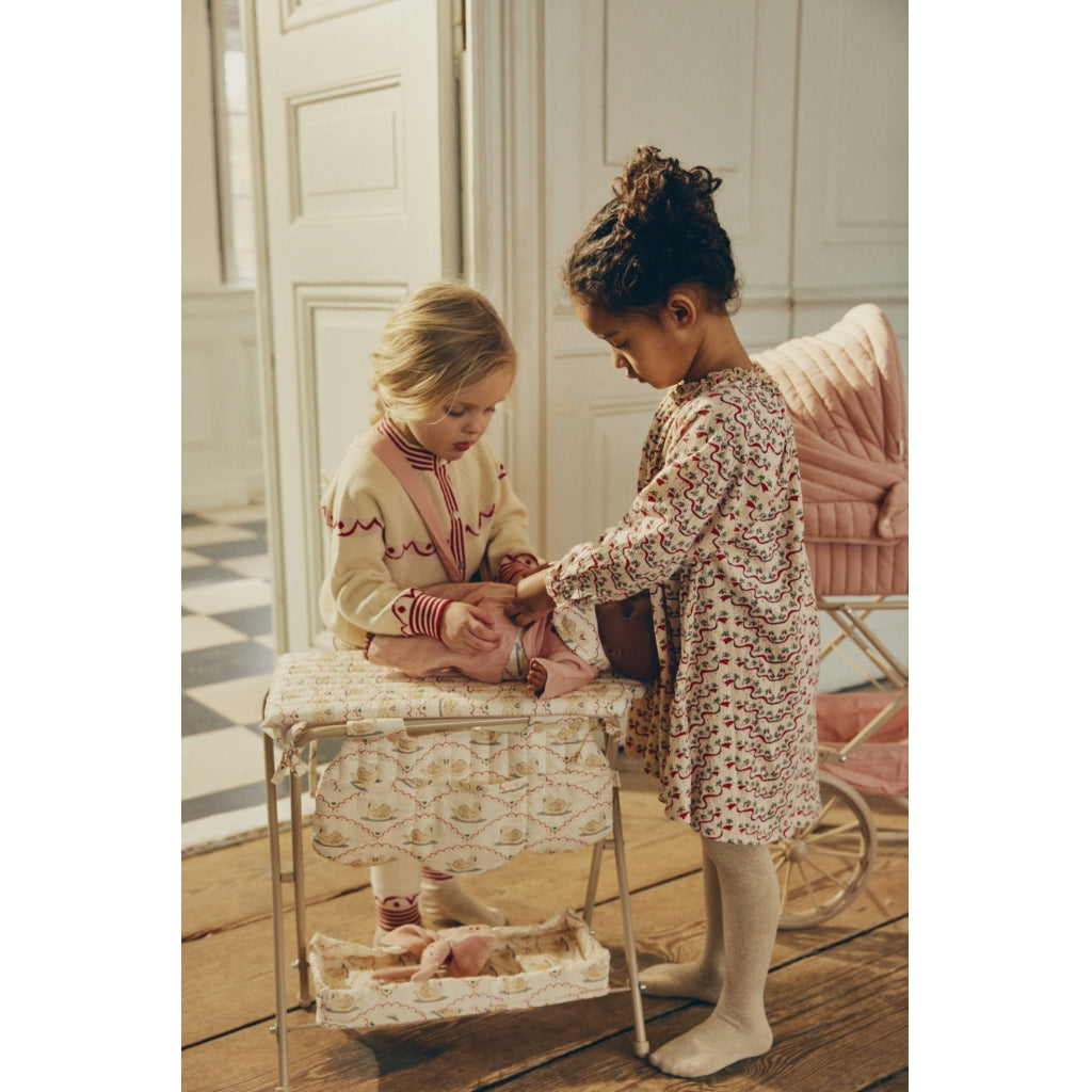 Two young girls play with a doll on a small changing table in a room with elegant wooden doors and a pink chair. One girl is helping to dress the doll in a Mahogany Rose doll pram.