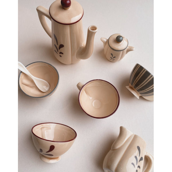 Sentence with product name: A collection of Konges Sløjd Pretend Play Porcelain Tea Set items in neutral tones with decorative elements, arranged on a light background, including a tall teapot, a smaller teapot, cups, and a creamer.