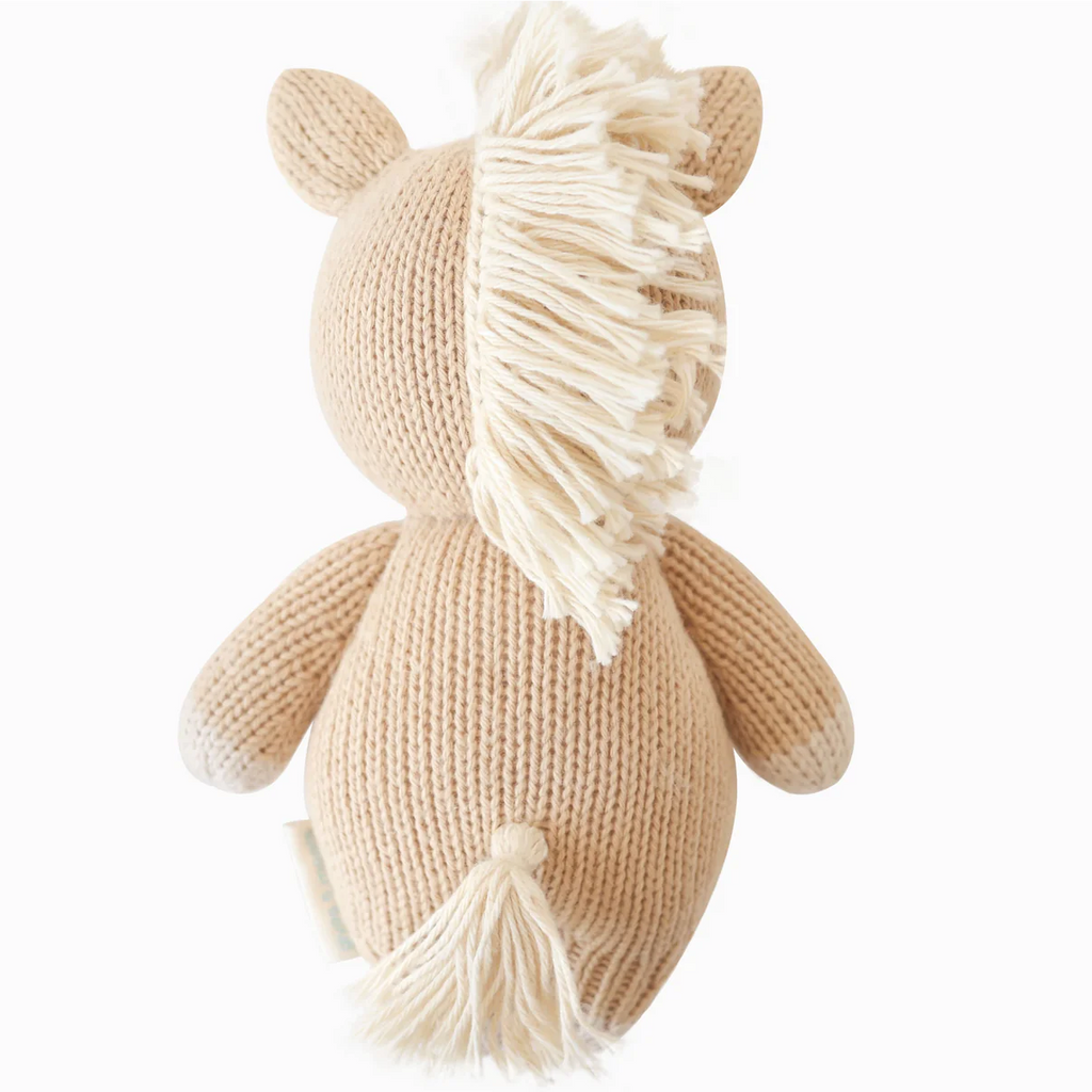 The Cuddle + Kind Baby Pony, hand-knit from natural Peruvian cotton yarn with a cream mane and tail, rounded body, and small ears, is shown from the back against a plain white background.