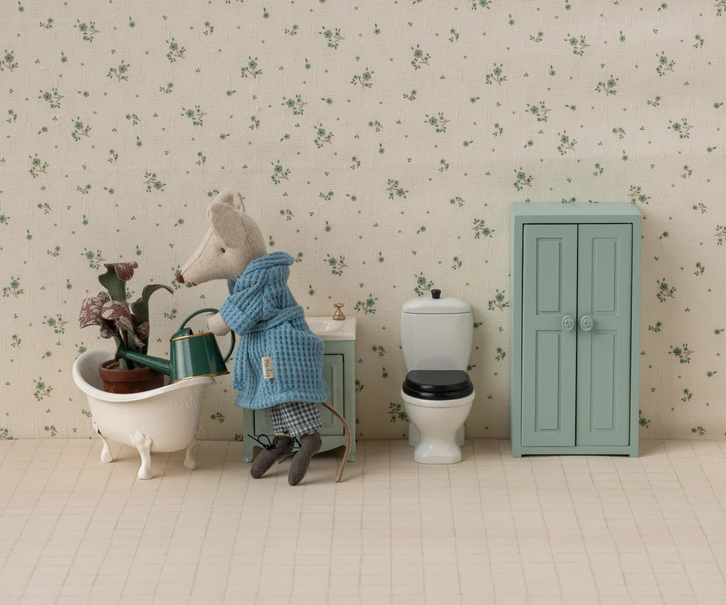 A mouse doll tends plants in a bathtub using the Maileg Watering Can, Mouse - Dark Green. The scene is a miniature bathroom with a toilet, light blue cabinet, and floral wallpaper.