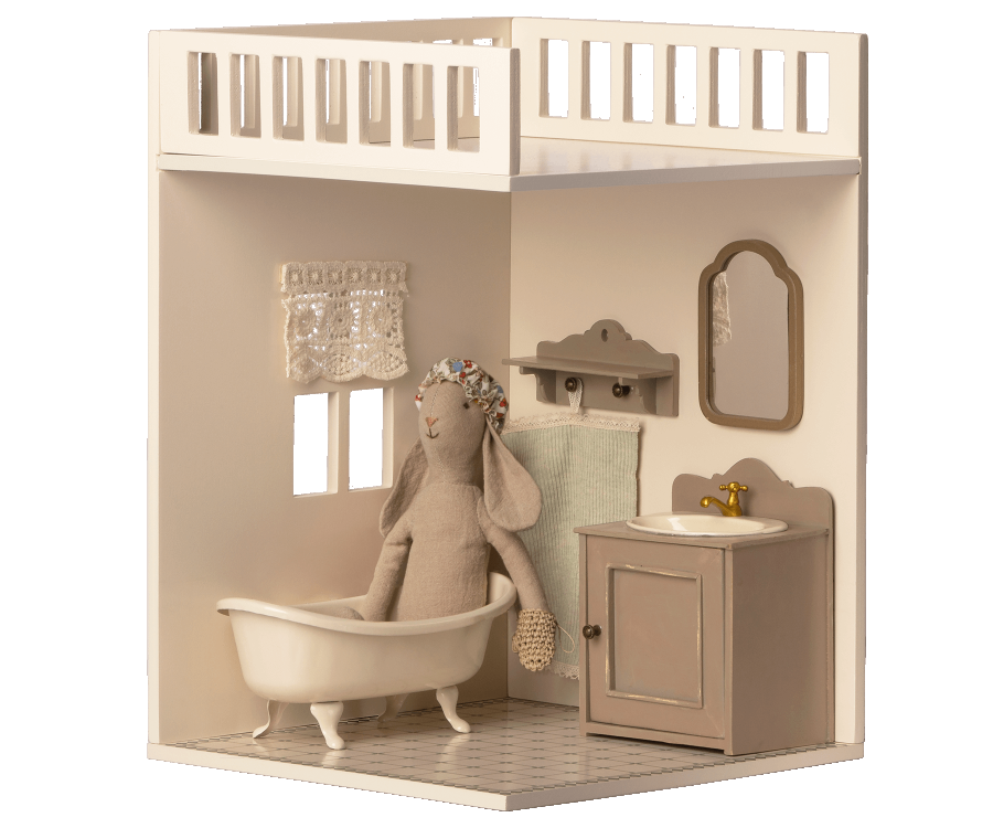 A plush rabbit toy wearing a shower cap sits in a mini bathtub in a Maileg House of Miniature Bonus Room - Bathroom, complete with a sink, mirror, and hanging towel.