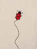 An embroidered red Ladybird with black spots and legs follows a dashed black line, set against a neutral textured organic cotton background.
