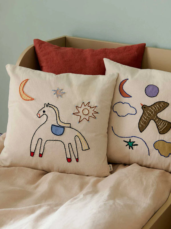 Two cream-colored Ferm Living Naïve Cushion - Horse with hand embroidered designs rest on a beige bed. The left pillow features a horse, crescent moon, and star. The right pillow shows two birds, clouds, and stars. The bed is partially covered with a light tan blanket, and a reddish-brown pillow is propped up in the back.