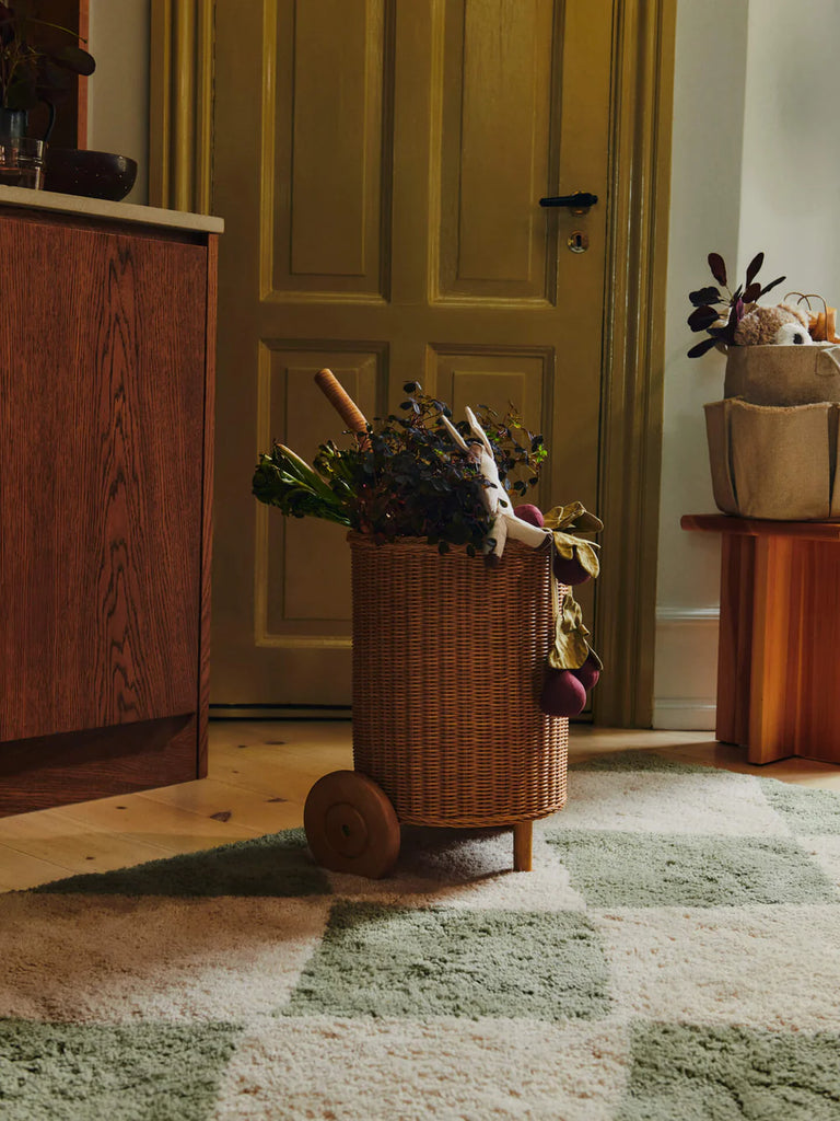 The Ferm Living Tukki Trolley - Natural, filled with greens and dried flowers, stands on a checkered rug in a cozy room featuring a yellow door and wooden furniture.
