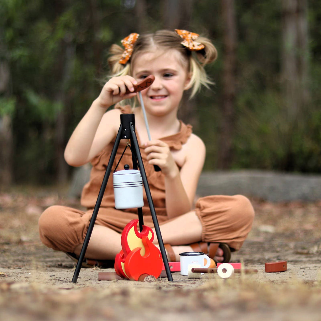 A young girl in brown overalls sits outside, happily pretending with the Wooden Camp Fire Set, which comes with a pot, tripod, and play food. She smiles as she holds a toy marshmallow on a stick over the wooden campfire.