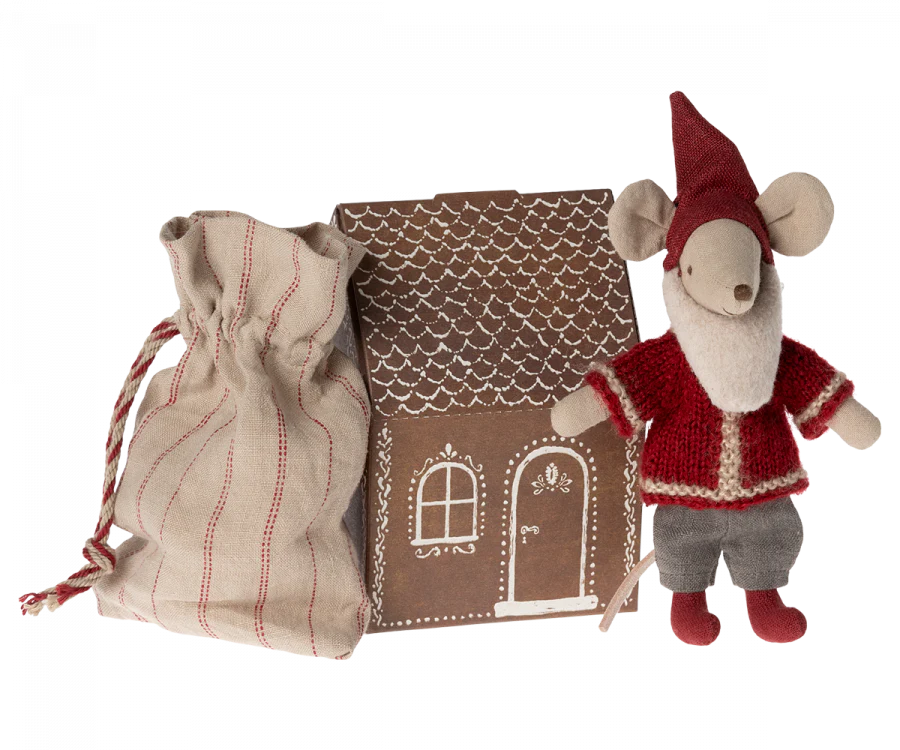 A small Maileg Santa Mouse, dressed in festive Santa clothes with a red hat, red and white outfit, and grey pants, stands next to a brown cardboard gingerbread house. The house features white icing-like decorations including a window and door. Nearby is a drawstring bag with red stitching—perfect for Christmas night.