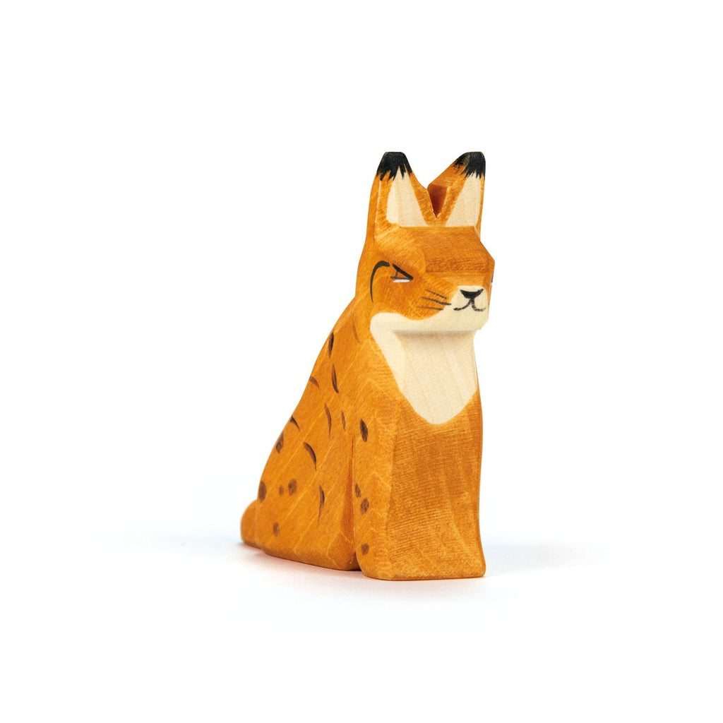 The Ostheimer Lynx Sitting is a small, hand-carved wooden figurine painted orange with dark spots and white chest and face accents, shown upright against a white background—ideal for imaginative play.