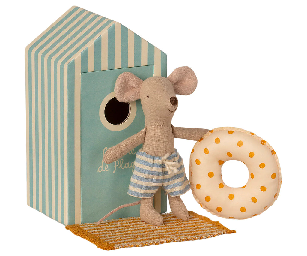 A small Maileg Beach Mouse - Little Brother wearing striped shorts holds a yellow polka-dotted inflatable ring. The mouse stands on a mat in front of a beach house with blue and white stripes, its circular entrance inviting vacation time.
