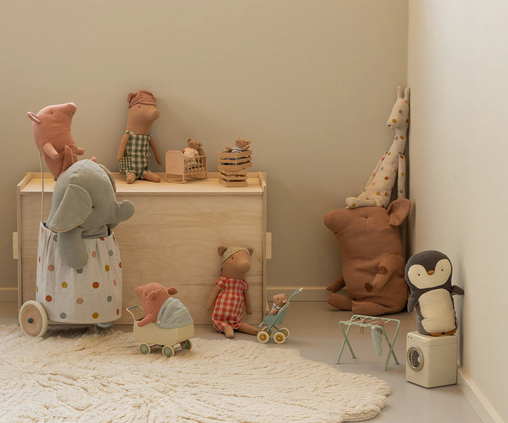 A cozy playroom corner features soft toys like the Maileg Truffle Pig, Medium, along with a penguin, elephants, bears, and a giraffe arranged around a wooden toy chest and small table on a light rug against neutral-colored walls.