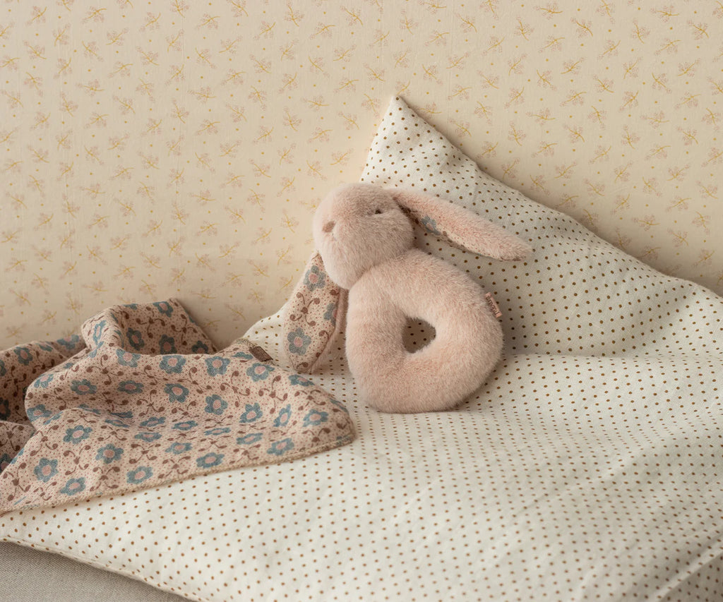 The Maileg Bunny Rattle Set - Powder includes a plush bunny on a polka-dotted pillow with a floral-patterned blanket, making it ideal for a charming gift. This cozy set is perfectly complemented by subtle star-patterned wallpaper in the background, creating an enchanting atmosphere.