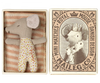 A plush toy dog with a neutral expression, wearing polka dot pants, lies inside a striped bed in an open box from the Maileg Farmhouse - Fully Furnished collection, with a decorative lid illustrating a royal canine character.