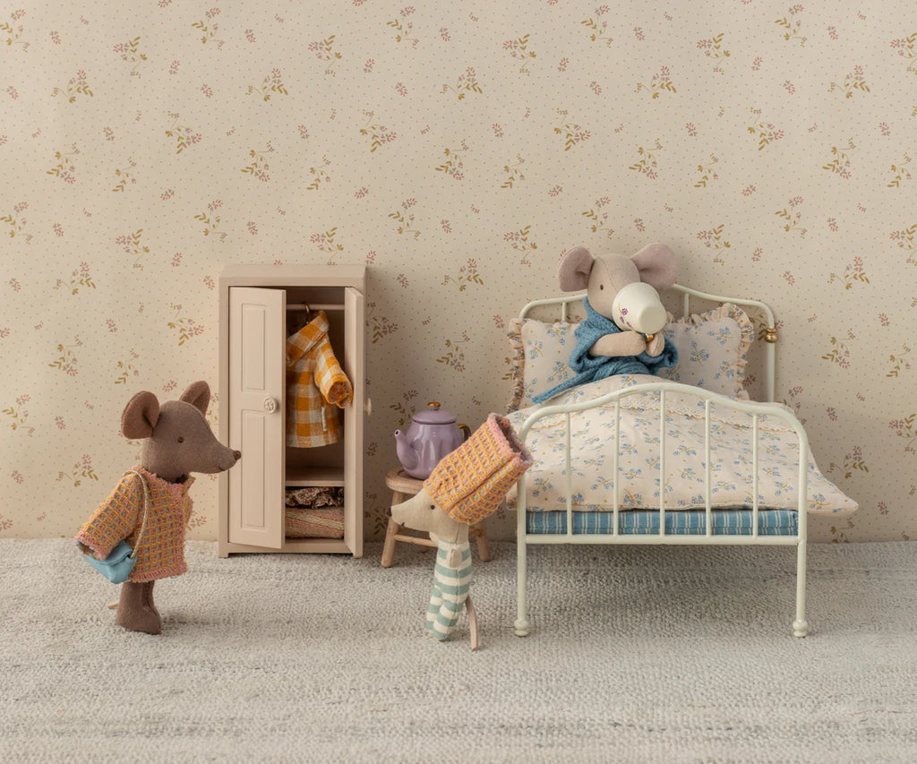 A cozy room with toy mice: one wearing a Maileg Suit for Mum Mouse sips from a cup on floral sheets, another stands with a blanket, and a third in a hat approaches. A small wardrobe and teapot complete the whimsical scene.
