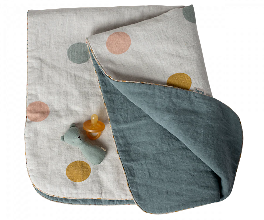 The Maileg Small Blanket - Chinos, shown light-colored with large pastel polka dots and a teal underside, is displayed with a green stuffed bear and yellow pacifier on a white background.