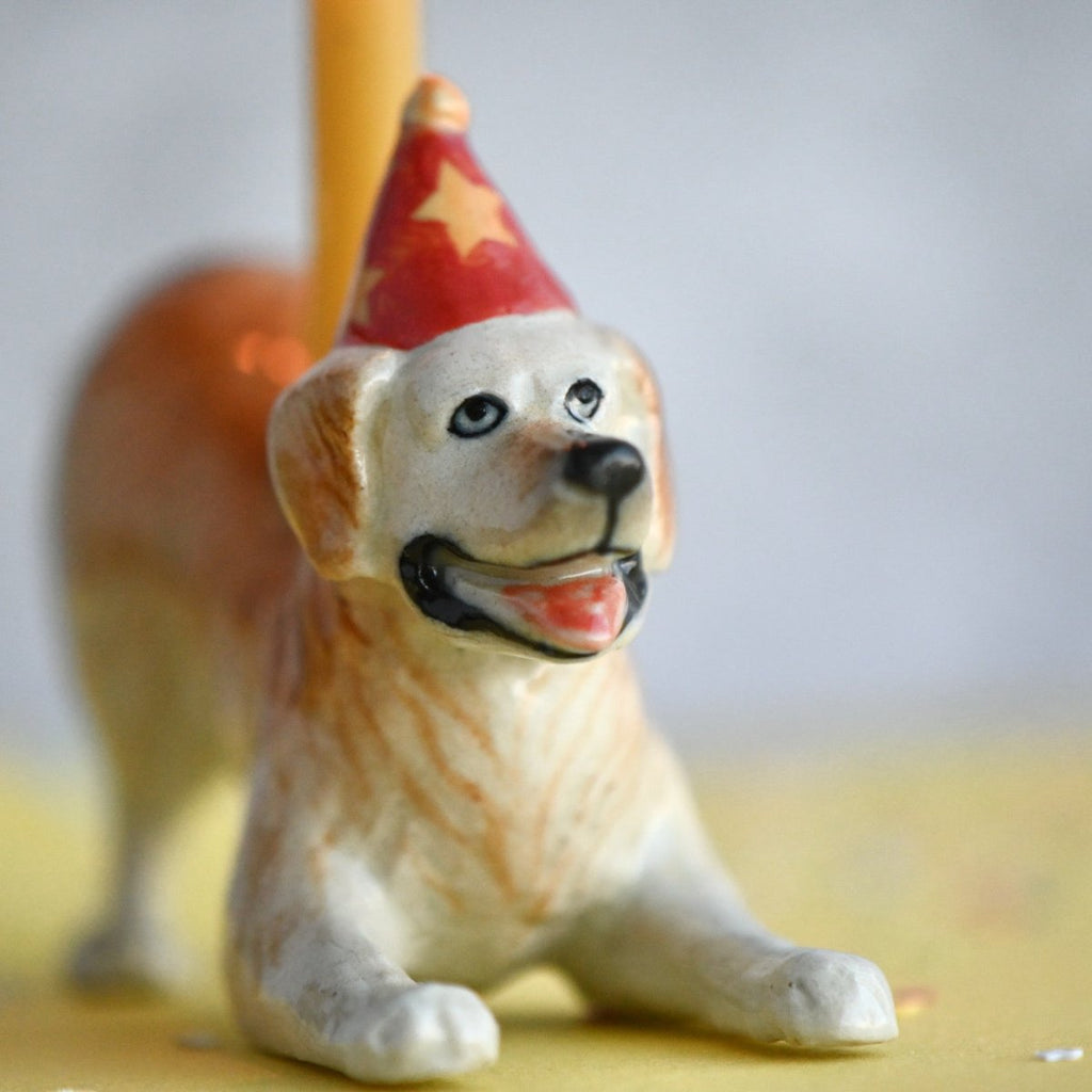 A close-up photo of a small hand painted Golden Retriever Cake Topper of a seated dog wearing a playful red and gold party hat, set against a soft, blurred background.