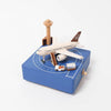 A model display of an Airport Music Box with a hand-cranked music box airplane, control tower, and baggage cart on a blue square base with white runway markings, crafted from sustainably sourced wood by.