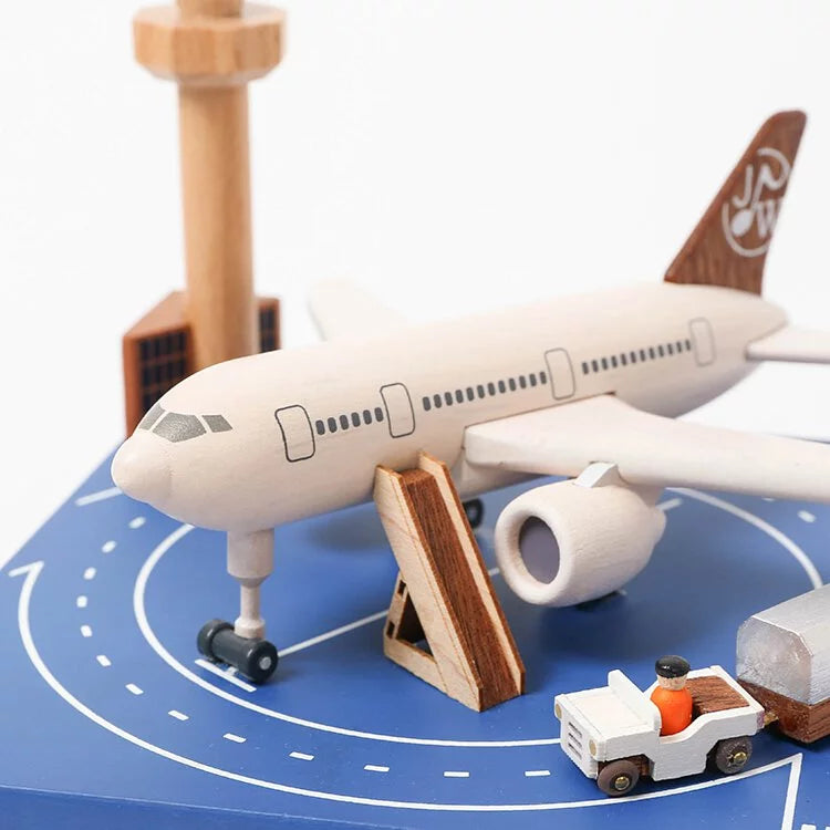 A model airplane on a runway mat surrounded by miniature airport elements including a control tower, baggage cart, and buildings, all crafted from sustainably sourced wood.