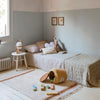 A cozy child's bedroom with a neat single bed, pastel blue walls, white carpet, and a small wooden stool. Toys are scattered near a Basket Honey Pot, and decorative pictures hang on the.