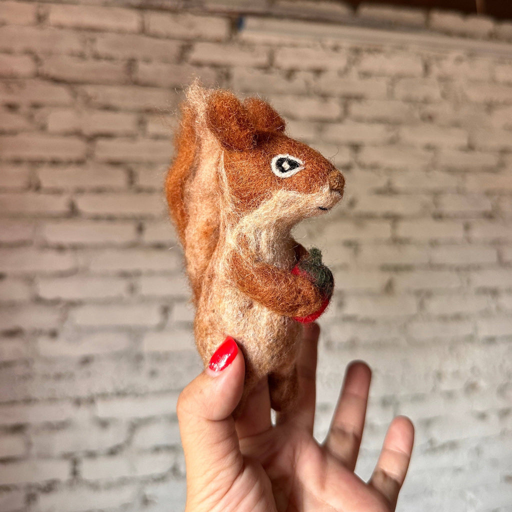 A hand with red nail polish holds the Squirrel Finger Puppet, a handmade wool accessory from Nepal, against a white brick wall. The small brown felt squirrel clutches a tiny red apple or berry.