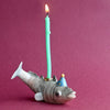 A whimsical hand-painted porcelain Fish Cake Topper shaped like a fish, wearing a party hat, with a lit birthday candle on its back against a pink background.