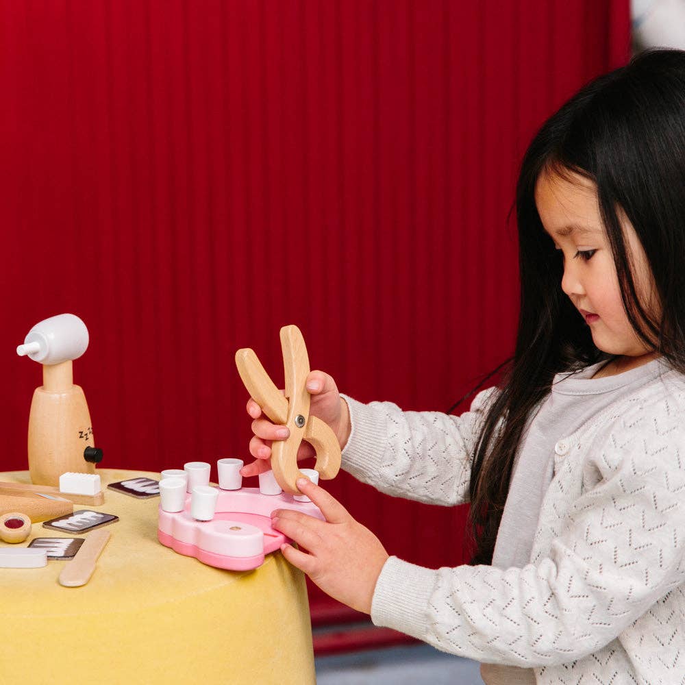 A child plays with the Wooden Dentist Set on a table, using large wooden tongs and exploring toy dental tools and teeth. A red curtain in the background creates an ideal setting for creative role play.