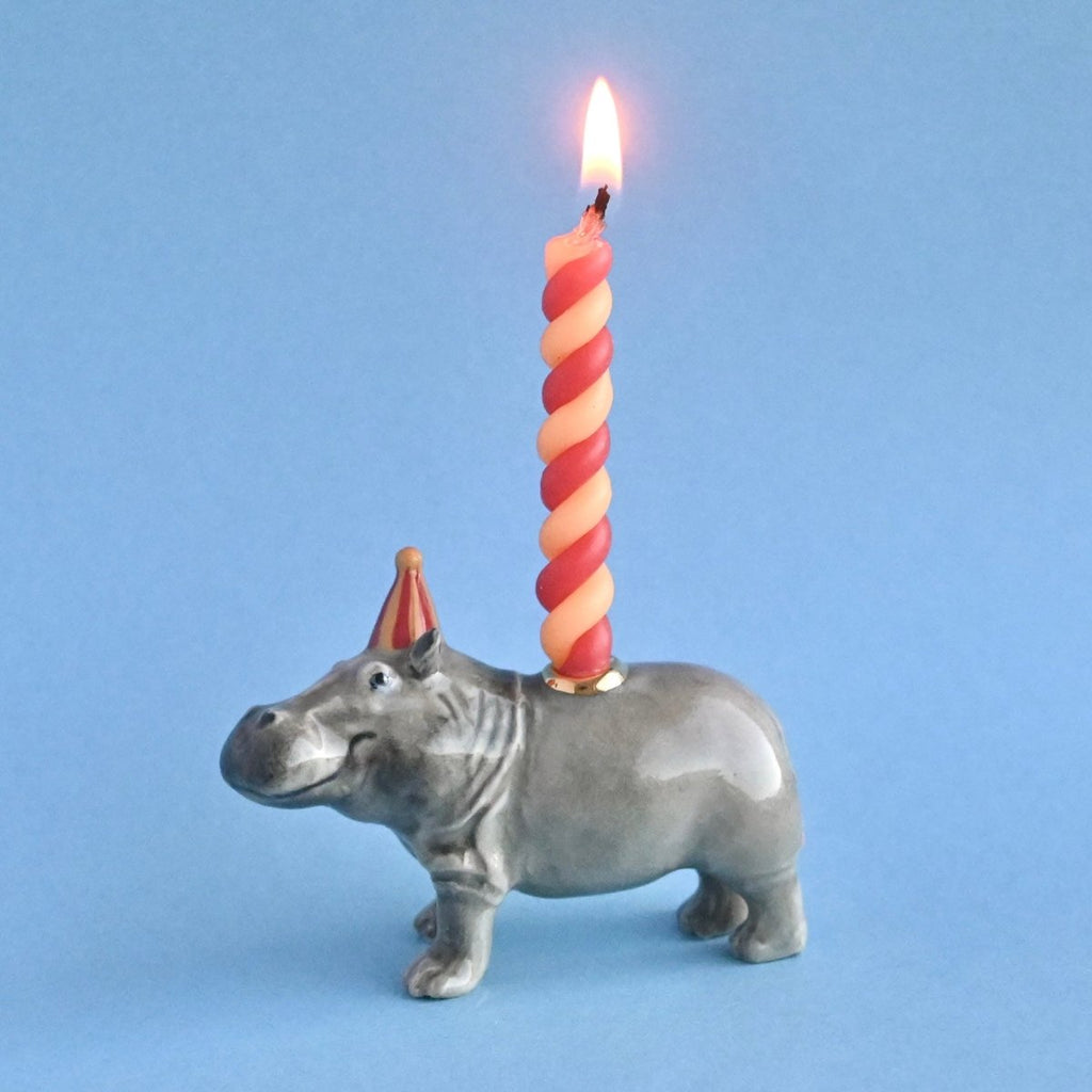 A fine porcelain Hippo Cake Topper with a red and white twisted candle lit on its back, set against a plain blue background.